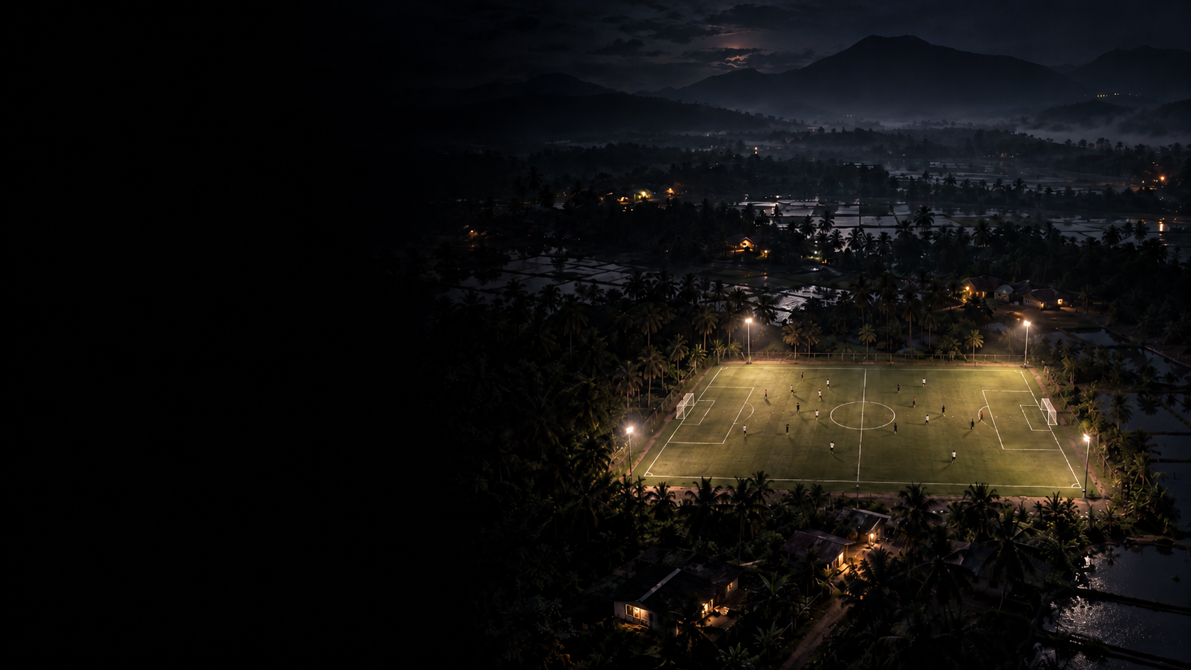 A floodlit football pitch at night, surrounded by palm trees and rice fields